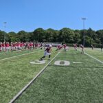 Football players warming up on a sunny field beside their team bench.